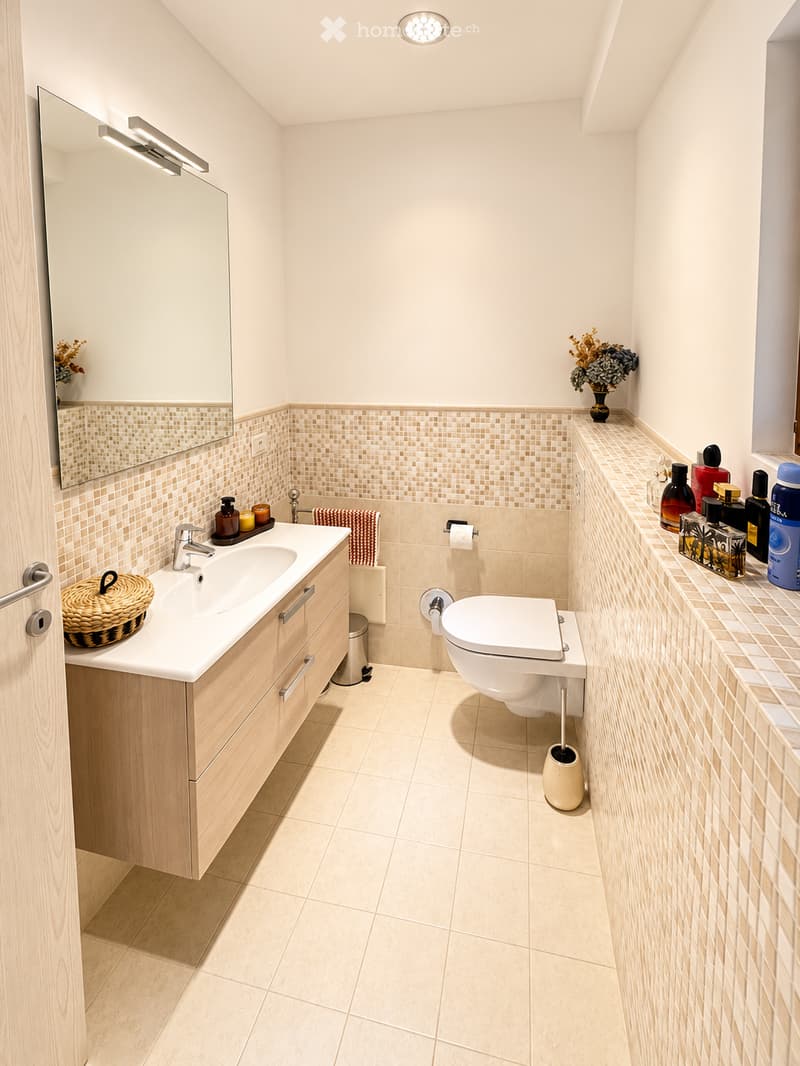 Modern bathroom with light-colored tiles, a wooden vanity, a sink, a toilet, a mirror, and decorative items including a wicker basket and a flower vase.