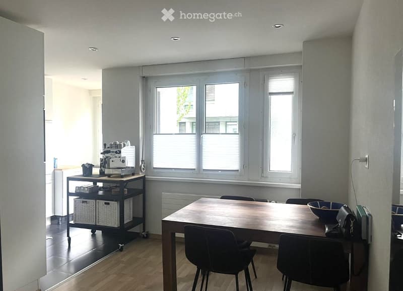 Bright room with wooden flooring, large windows, a dining table with chairs, a coffee machine on a cart, and a bowl on the table.