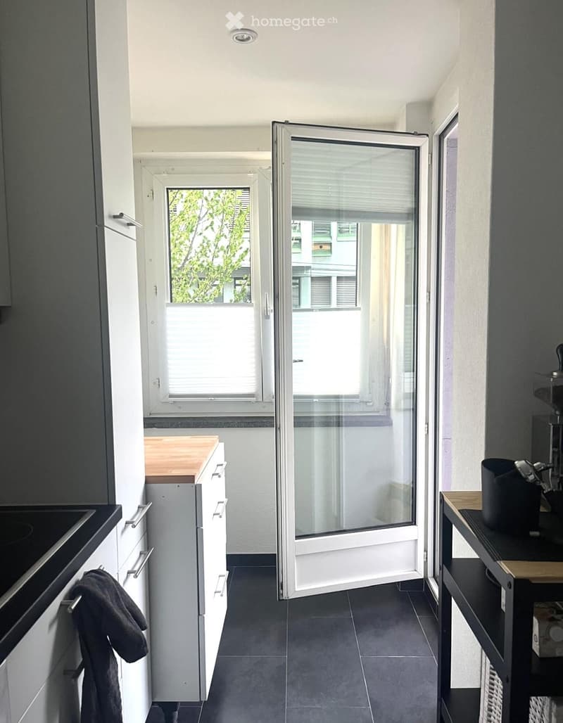 Modern kitchen with white cabinets, dark countertops, and a sliding glass door leading to a balcony. Includes a refrigerator, sink, coffee maker, and a small table.