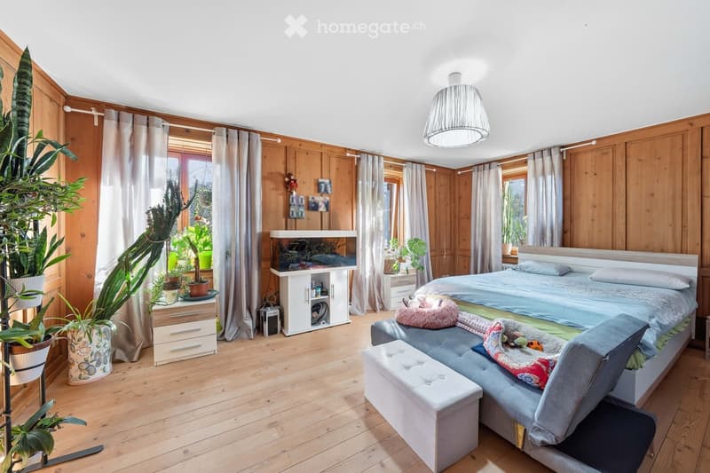 Cozy bedroom with wooden walls and flooring, featuring a large bed, a sofa, a stool, a nightstand, a fish tank, a drawer, and several potted plants.