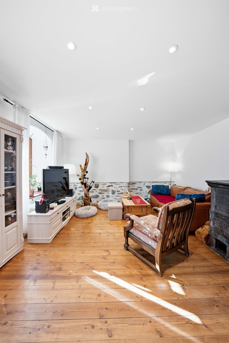 Bright living room with wooden flooring, white walls, and a mix of modern and rustic furniture including a sofa, armchair, coffee table, TV, and fireplace.
