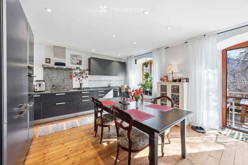 Modern kitchen with dark grey cabinets, stainless steel appliances, a wooden dining table with four chairs, and a view of a snowy outdoor area through large windows.