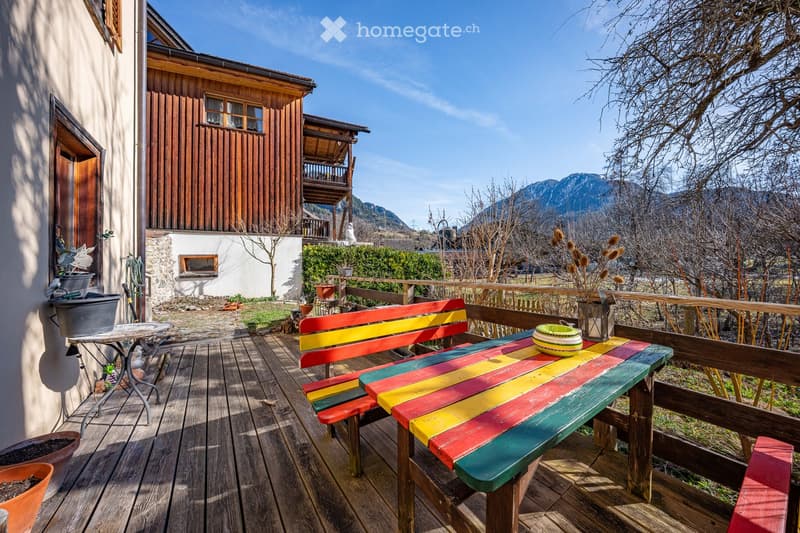 Wooden balcony with colorful bench and table, overlooking a scenic mountain view with surrounding greenery and a wooden house.