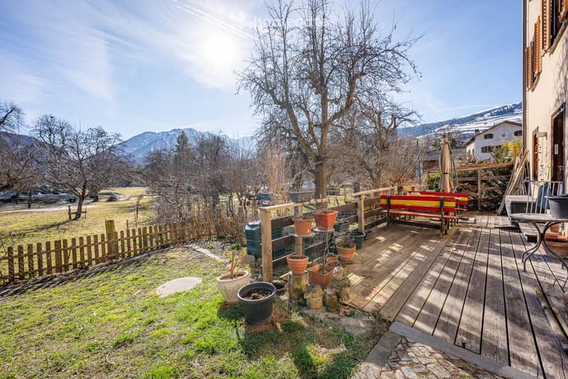 A wooden deck with a bench, potted plants, and a view of a grassy area with trees and mountains in the background.