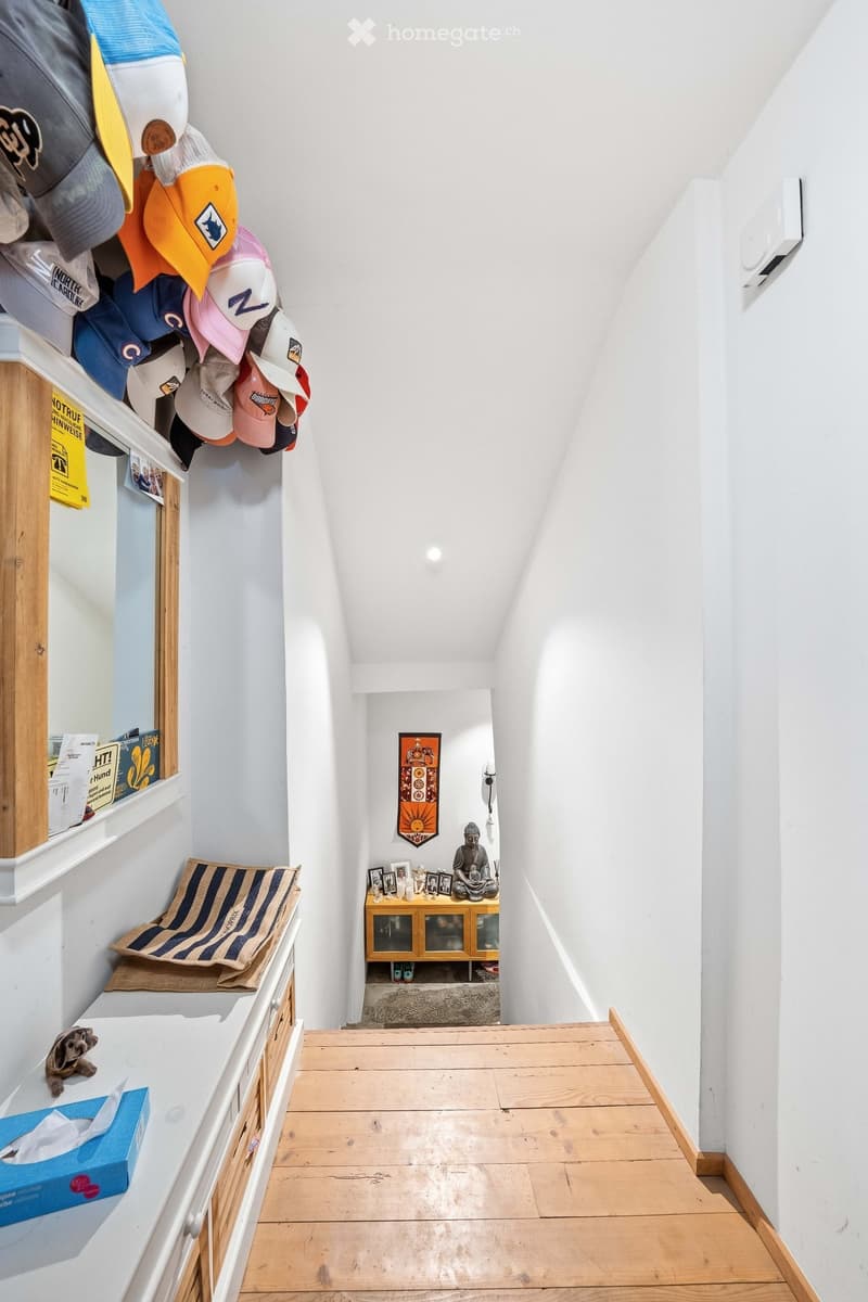 A narrow hallway with wooden flooring, white walls, and a small shelf displaying hats and other items. A small table with a statue and framed photos is visible at the end of the hallway.
