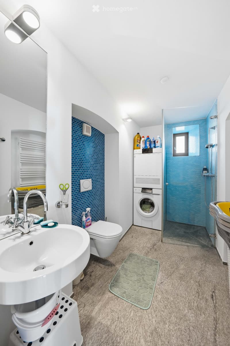Modern bathroom with a sink, toilet, shower, washing machine, and cleaning supplies on a shelf. Features a blue tiled accent wall and a green bath mat.