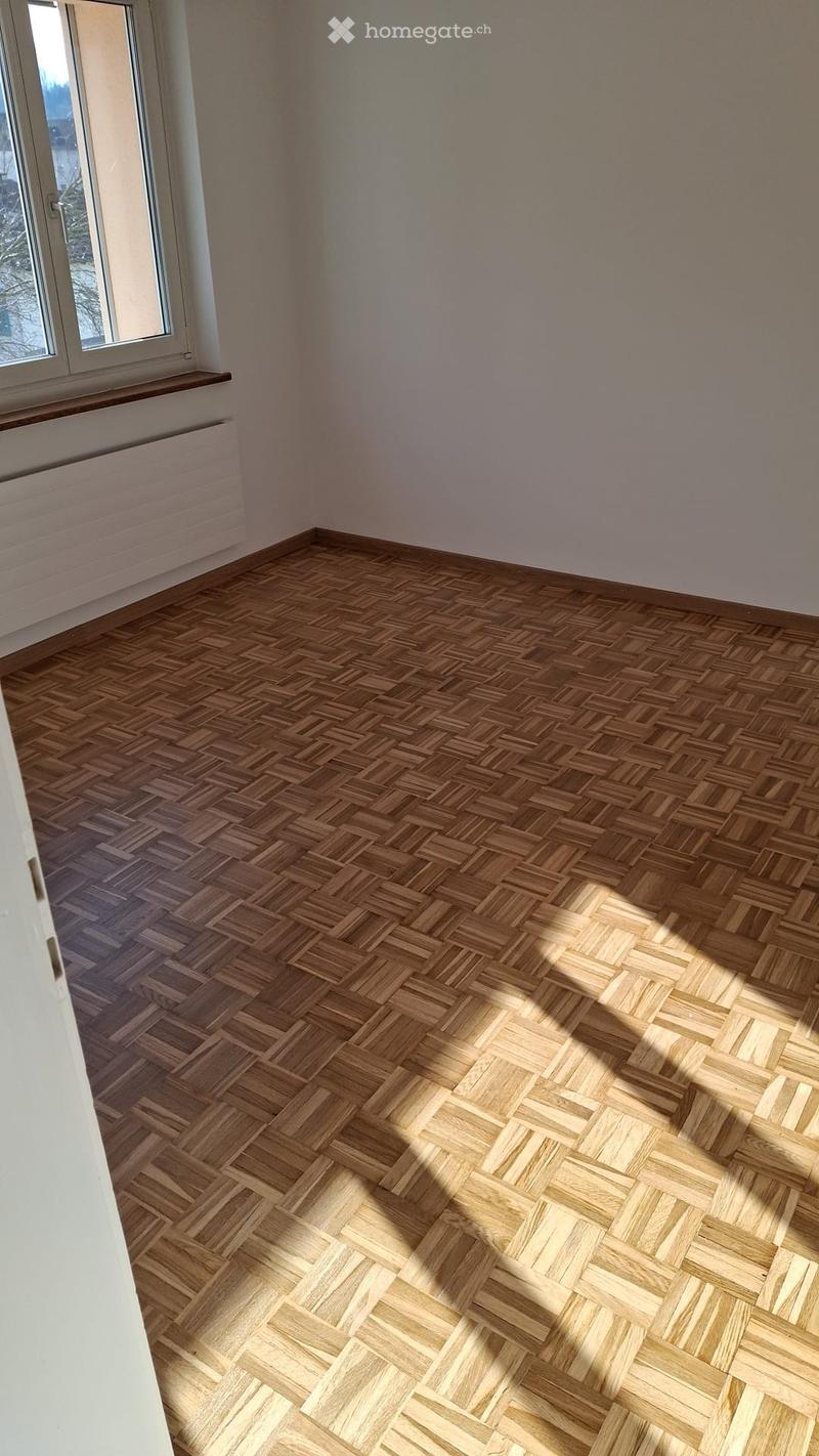 Empty room with herringbone-patterned wooden flooring and white walls, featuring a large window with a radiator below it.