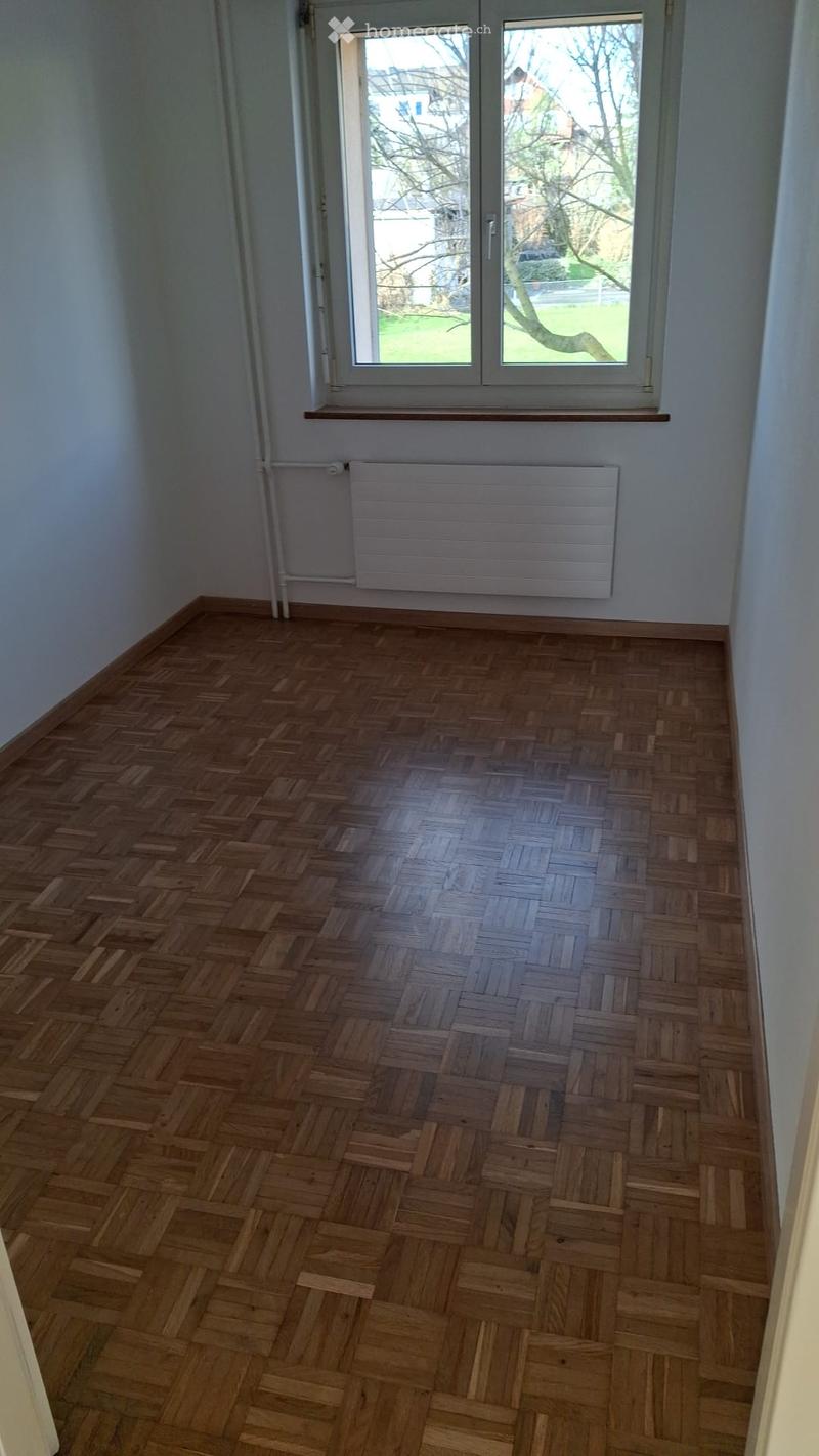 Empty room with wooden herringbone flooring, white walls, a radiator, and a large window with a view of a garden.