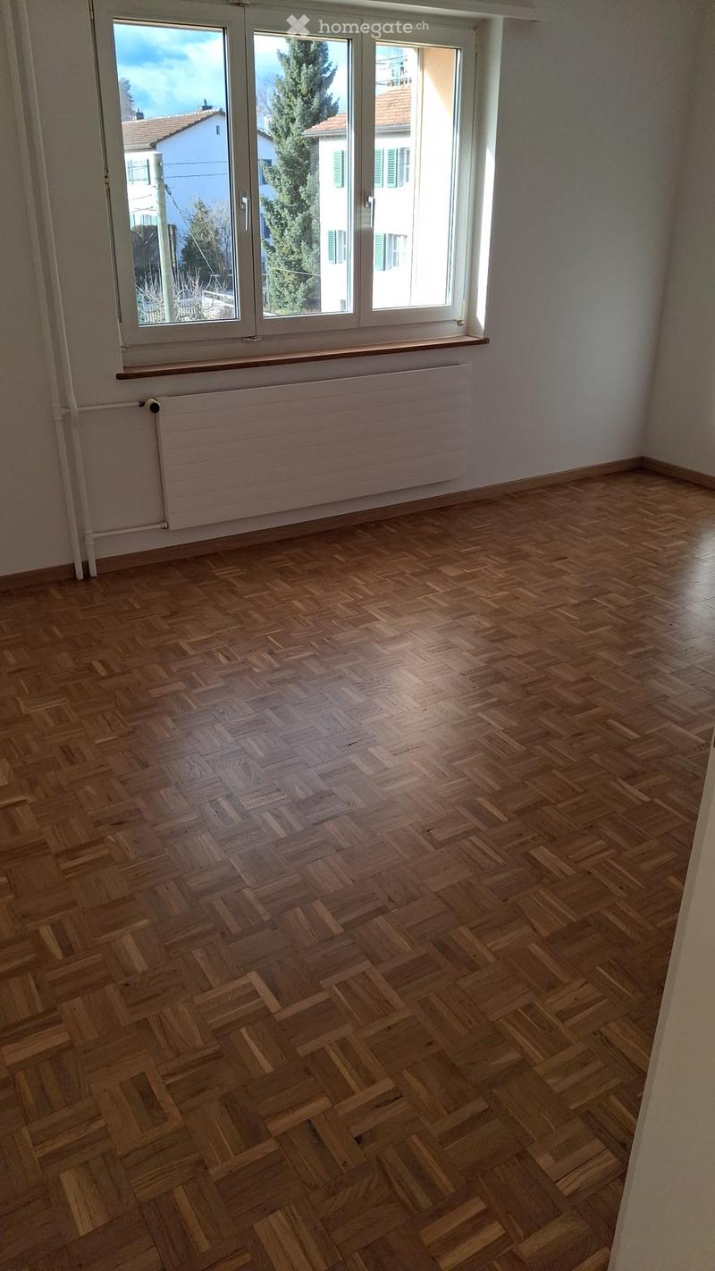 Empty room with wooden herringbone flooring, large windows with white frames, radiator, and a small wooden shelf above the radiator.