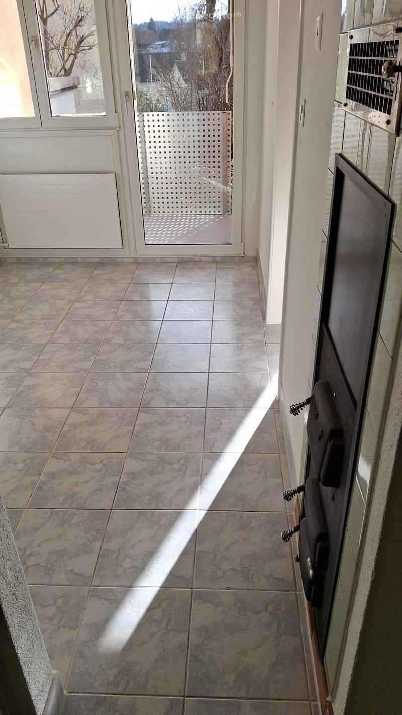 Empty hallway with tiled floor, white walls, and a glass door leading outside. Natural light streams in from the left side.