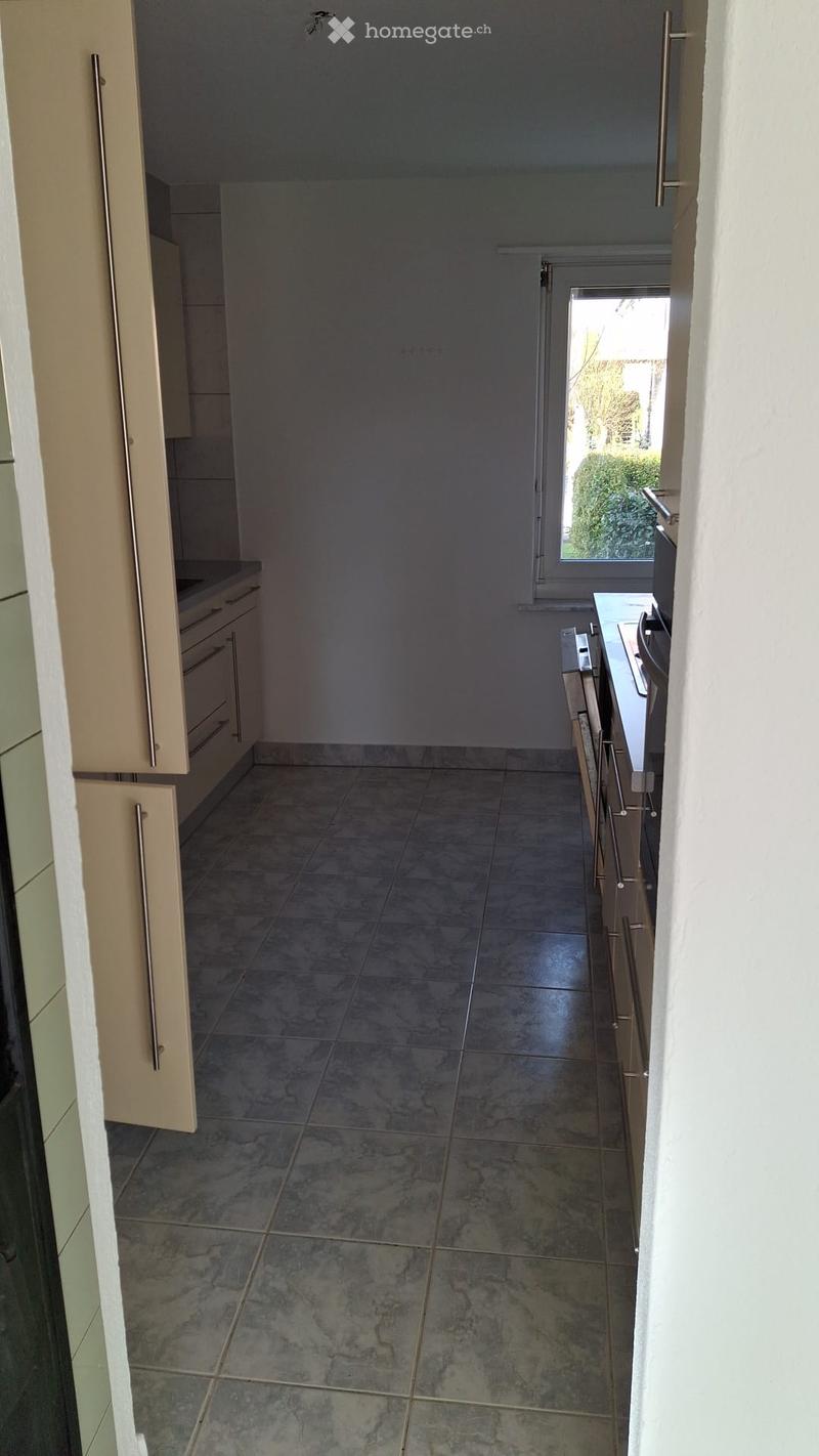 Empty kitchen with light-colored cabinets, tiled floor, and a window overlooking a garden area.