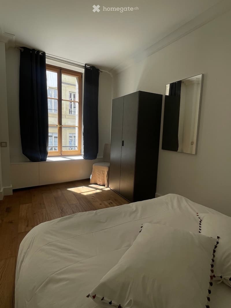 Bright bedroom with wooden flooring, large window with black curtains, bed with white duvet, wardrobe, and a small stool near the window.