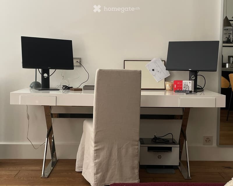 A modern, minimalist workspace with a white desk, two computer screens, a chair, and a small computer tower under the desk.
