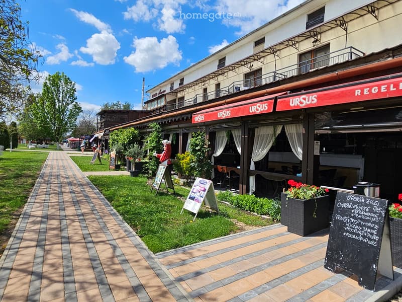 A restaurant named "URSUS" with outdoor seating, a red awning, and a well-maintained garden area with potted plants and a menu board.