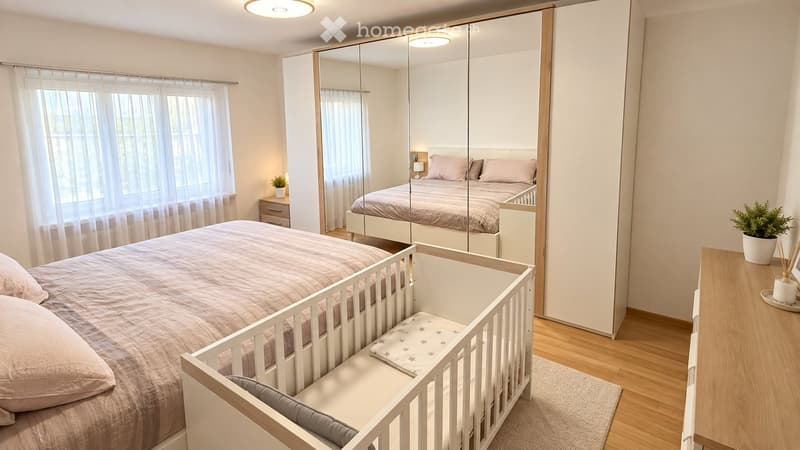 Bright bedroom with a large bed, mirrored wardrobe, nightstand, baby crib, and potted plants. The room features light-colored walls, wooden flooring, and large windows with sheer curtains.
