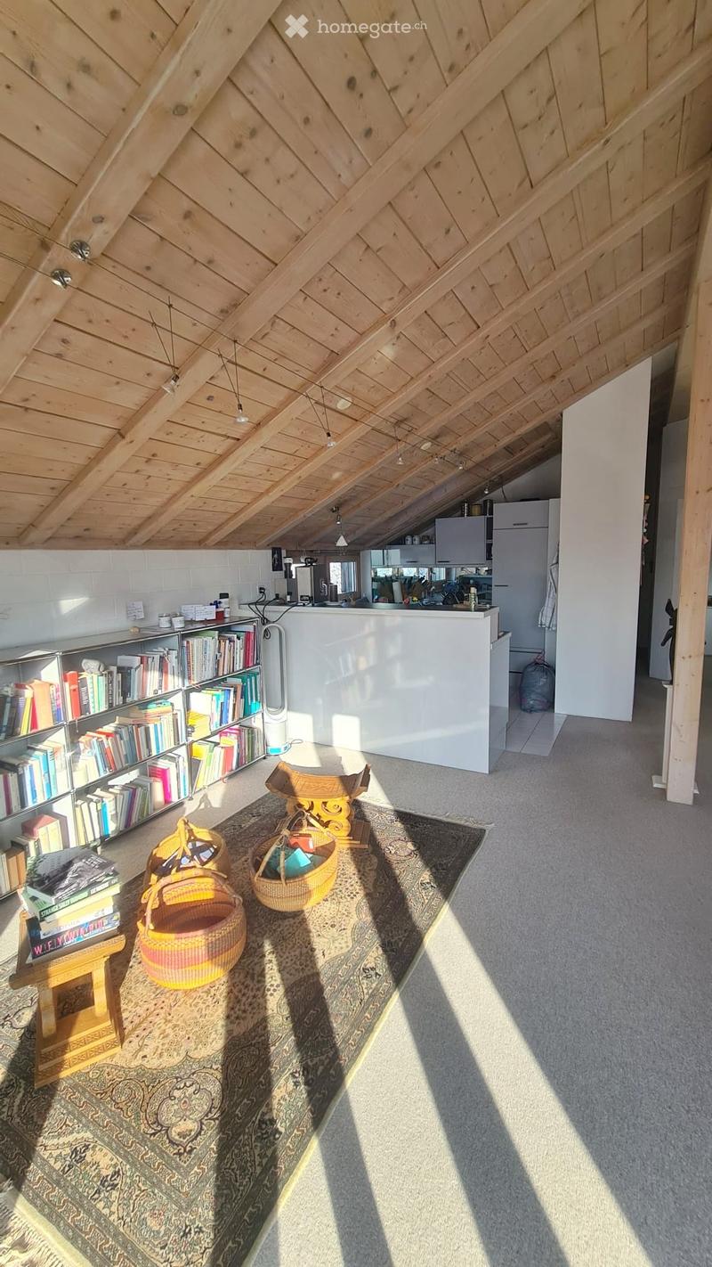 Bright, open living space with wooden slanted ceiling, bookshelf, small table, woven baskets, and a modern kitchen area in the background.