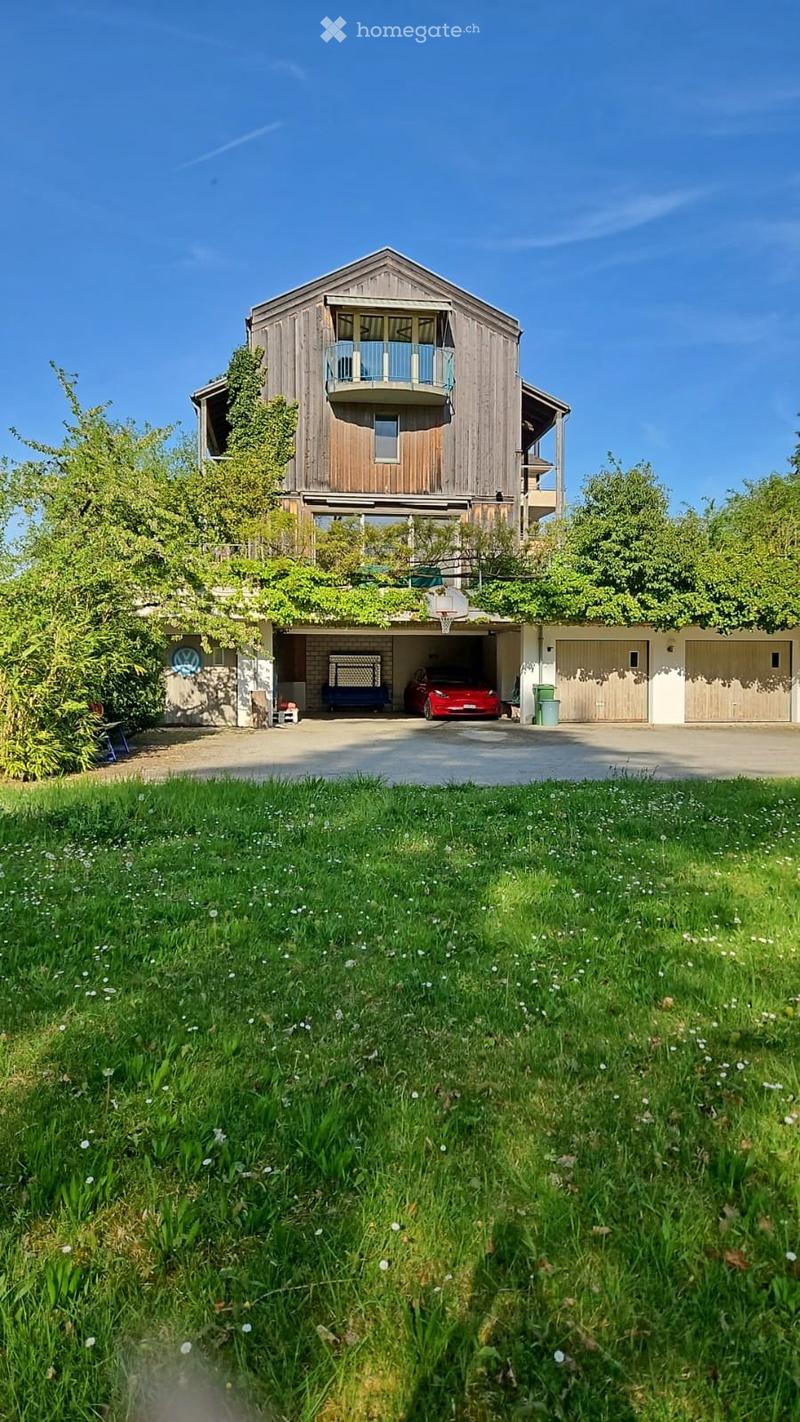 A modern two-story house with a wooden exterior, a balcony on the upper level, and a large garage with multiple parking spaces. The house is surrounded by a well-maintained lawn and trees.