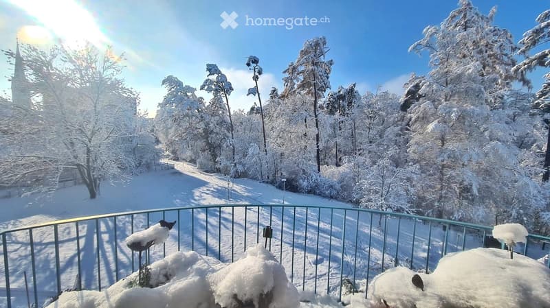 A snowy outdoor balcony overlooking a snow-covered forest and a distant church spire, with a metal railing and snow-covered furniture.