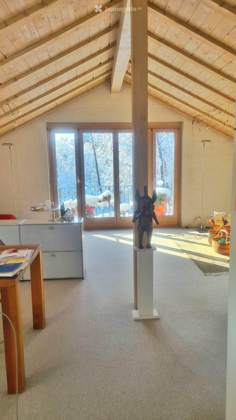 Bright, open attic space with wooden beams, large windows, and a view of snow-covered trees. Includes a table, refrigerator, statue on a pedestal, floor lamp, and wicker baskets.