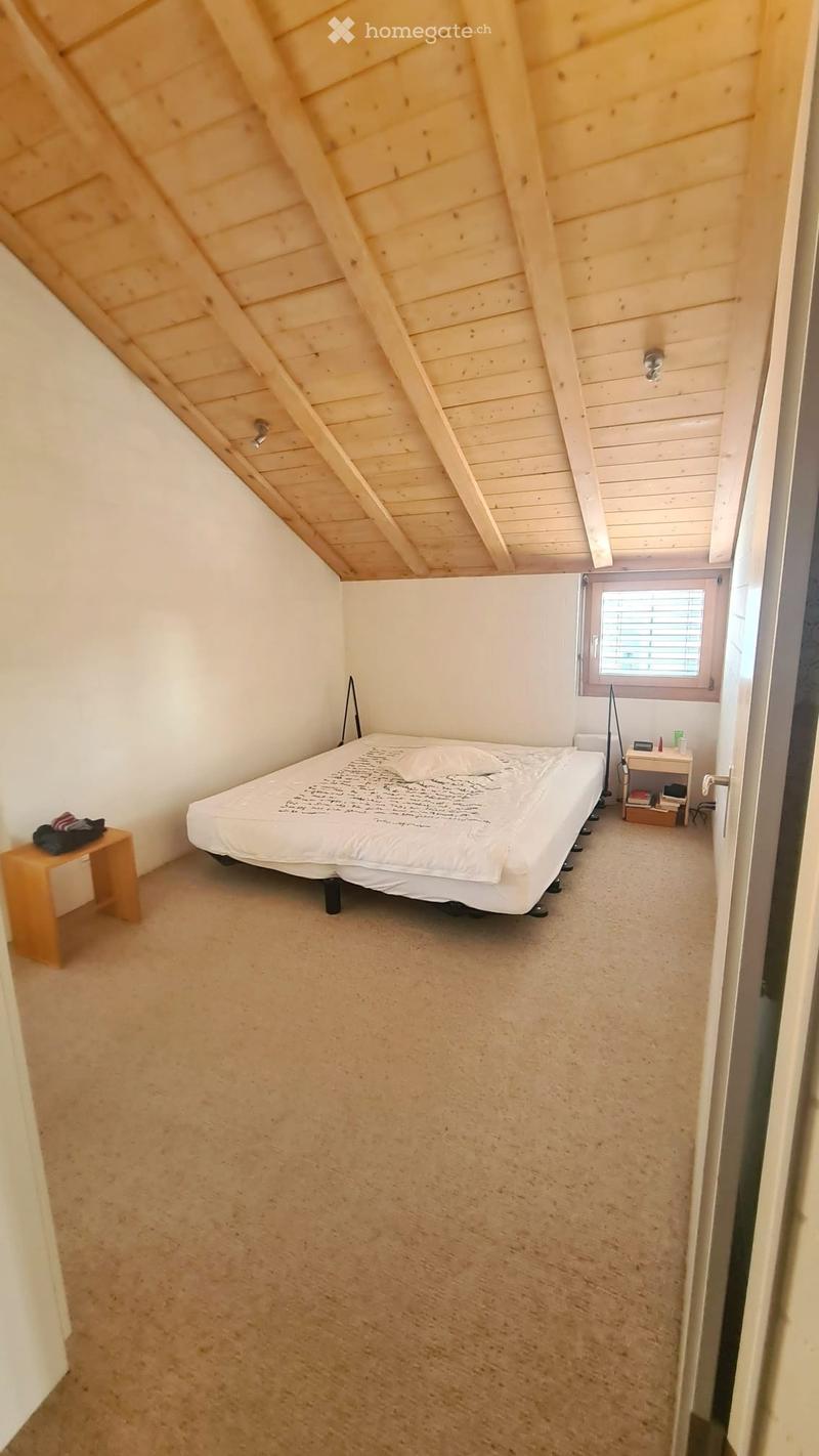 Cozy attic bedroom with a slanted wooden ceiling, a single bed, a small nightstand, and a chair. The room has carpeted flooring and a window with blinds.