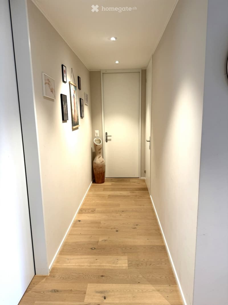 Modern hallway with light wooden flooring, white walls, recessed ceiling lights, and framed artwork. A tall decorative vase is placed near the door.