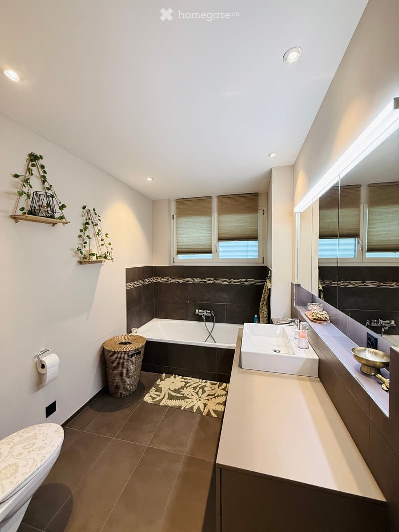 Modern bathroom with a bathtub, sink, toilet, and large mirror. Features dark brown tiles, white walls, and a decorative plant on a shelf.