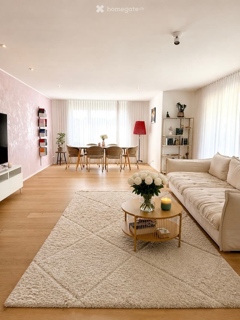 Modern living room with light wooden flooring, white walls, and large windows covered by sheer curtains. Features a cream-colored sofa, a round coffee table with a vase of white flowers, a dining area with a table and chairs, a floor lamp, and a shelf wit