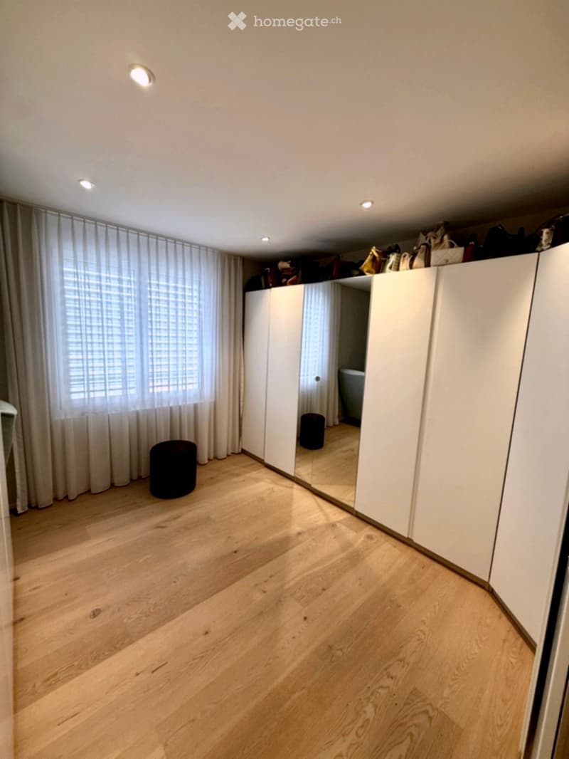 Empty room with wooden flooring, large window with white curtains, white closet, and a small black stool near the window.