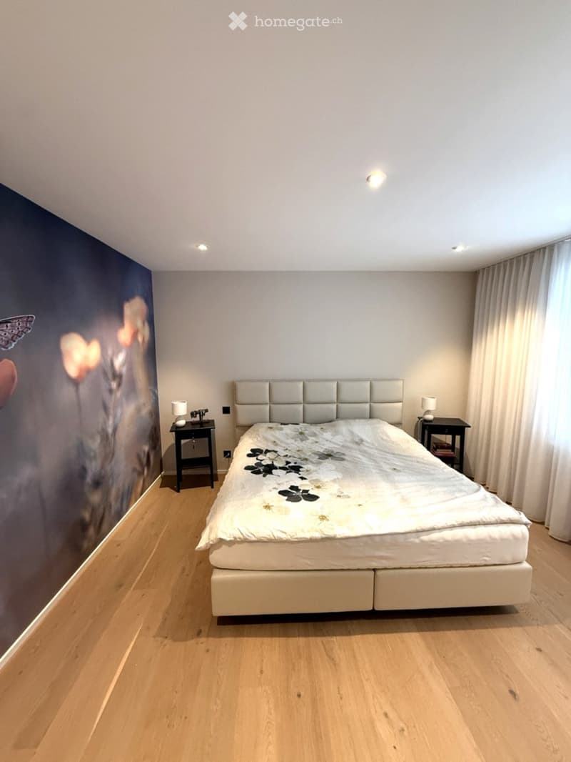 Modern bedroom with wooden flooring, large bed with white headboard and floral duvet, two nightstands with lamps, large window with curtains, and a mural on one wall.