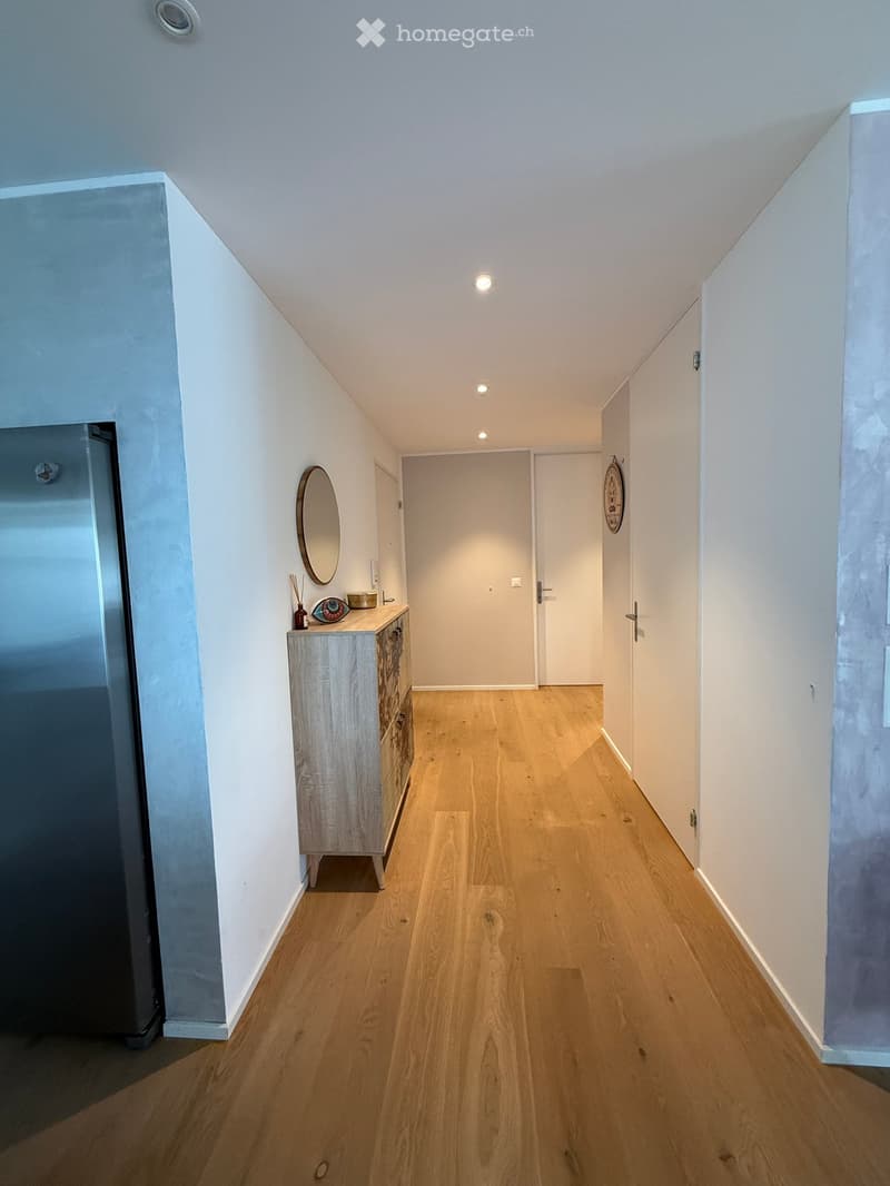 A narrow hallway with wooden flooring, white walls, and recessed ceiling lights. A small wooden cabinet with drawers and a mirror is positioned on the left side, with a door at the end of the hallway.