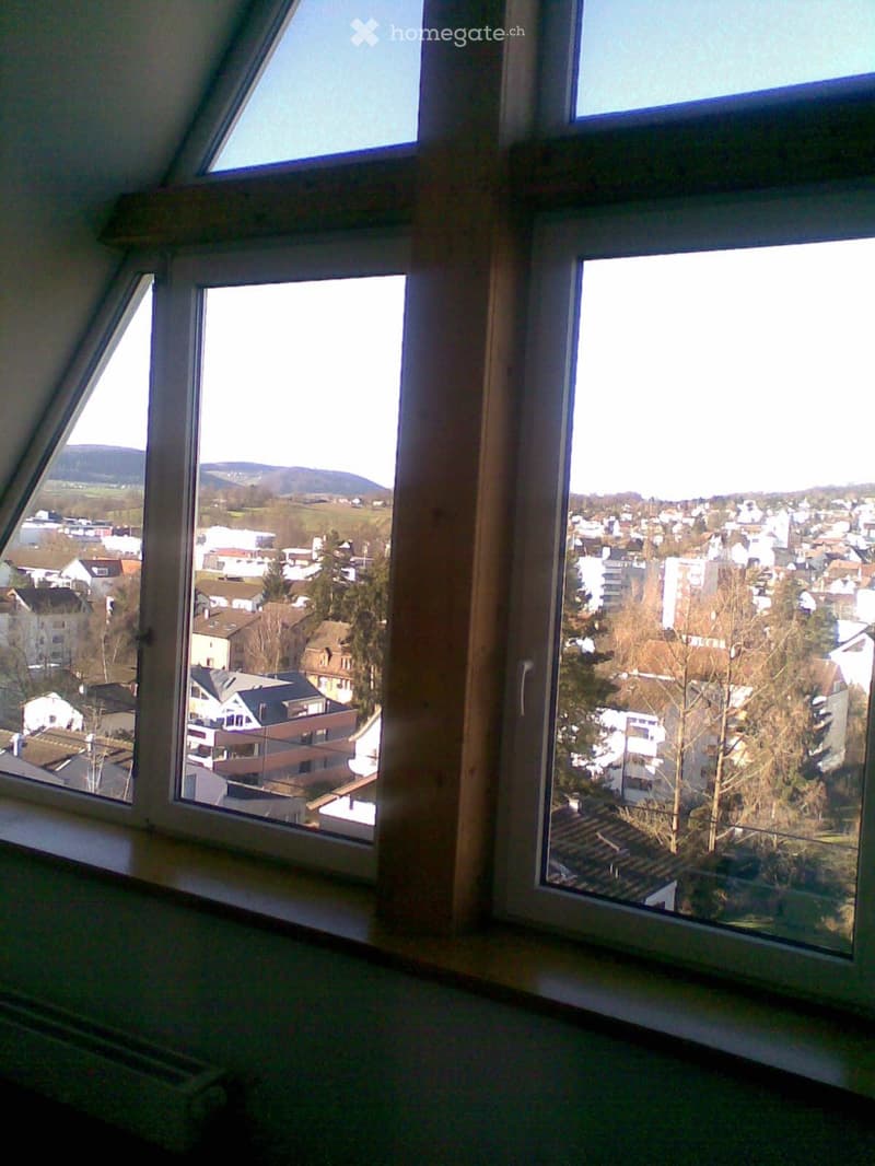 An unfurnished room with large windows offering a panoramic view of a town and surrounding landscape, including houses, trees, and distant hills.