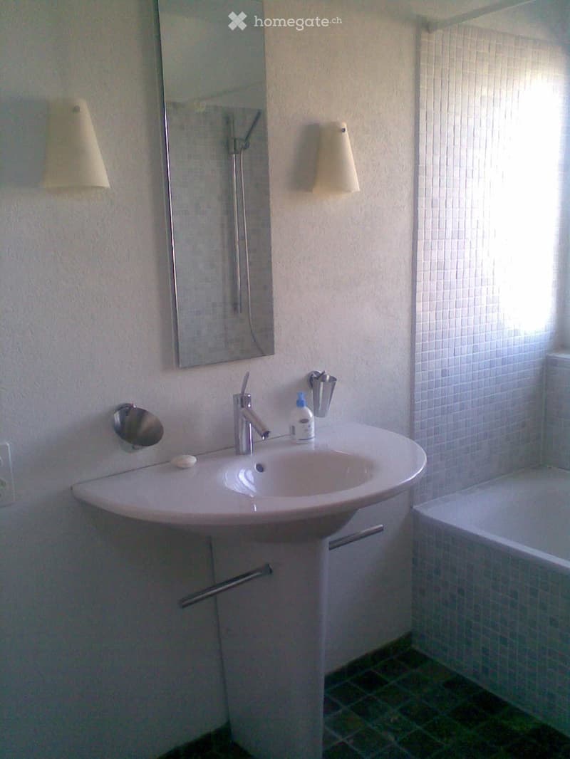 Modern bathroom with a white sink, chrome faucet, mirror, and towel racks. The sink has a sleek, curved design, and the walls are tiled with light-colored tiles.