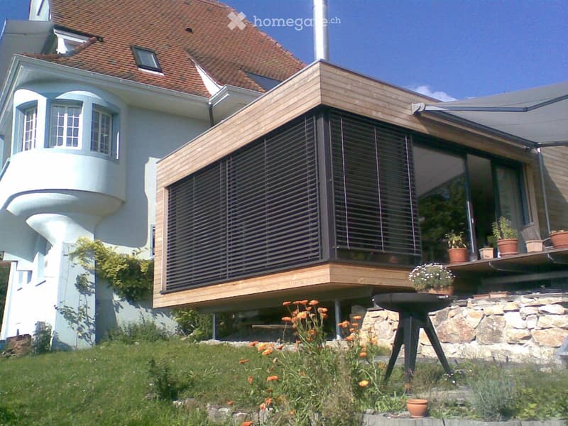 Modern two-story house with a curved facade, large windows, and a raised ground floor. Features include a balcony with potted plants, a stone staircase, and a well-maintained garden with orange flowers.