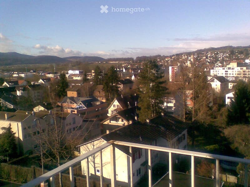 View from a balcony showing a suburban neighborhood with houses, trees, and distant hills under a partly cloudy sky.