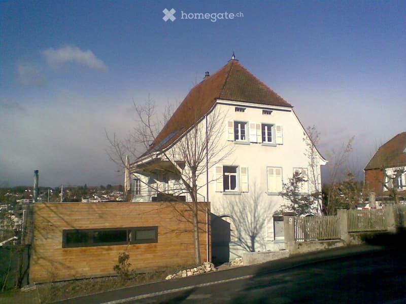 Two-story house with white exterior, red-tiled roof, multiple windows with shutters, and a wooden garage section.
