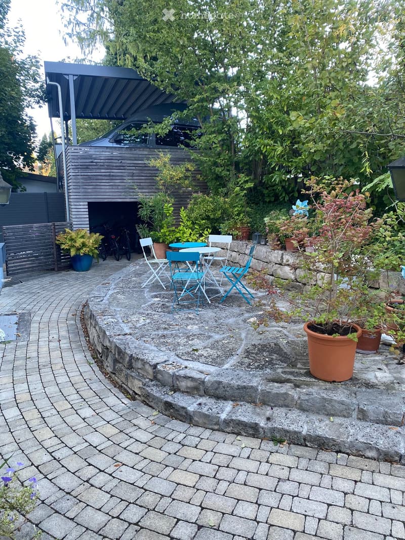 Garden area with a stone patio, a small seating area with a table and chairs, potted plants, and a garage in the background.