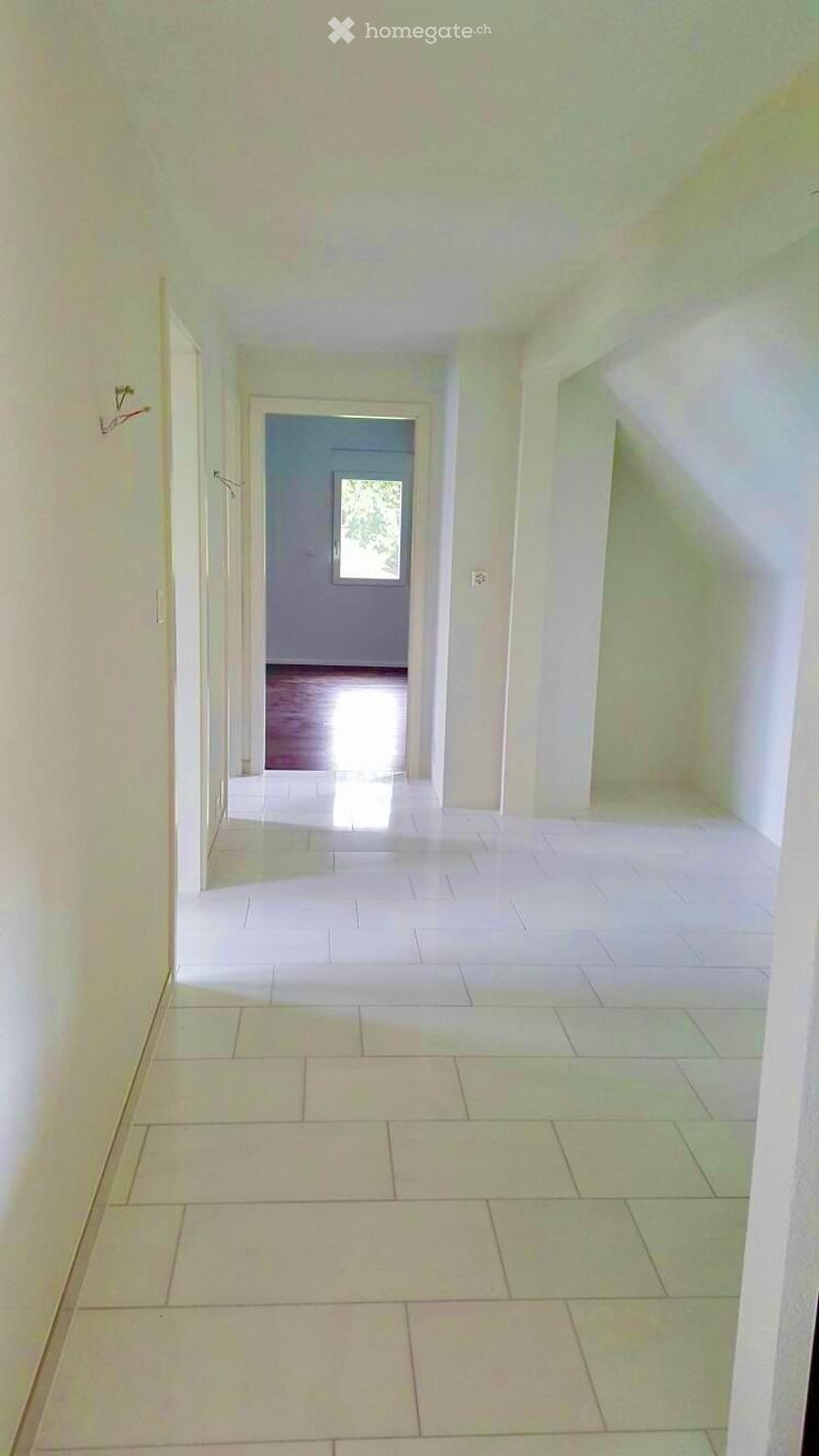 Empty hallway with white tiled flooring and light-colored walls, leading to a room with wooden flooring and a window.