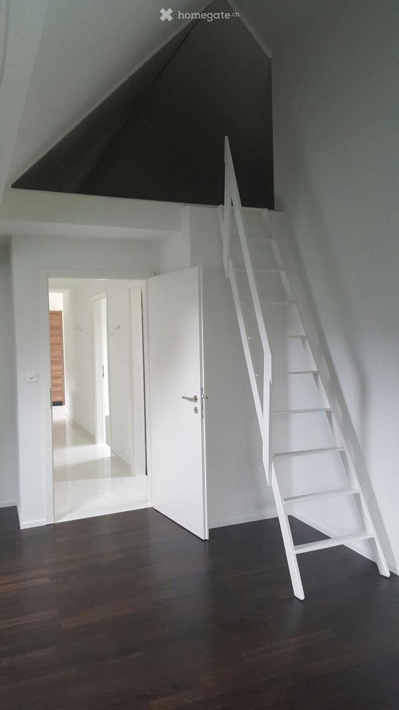 Empty attic space with wooden flooring, white walls, and a white staircase leading upwards. An open doorway leads to another room.