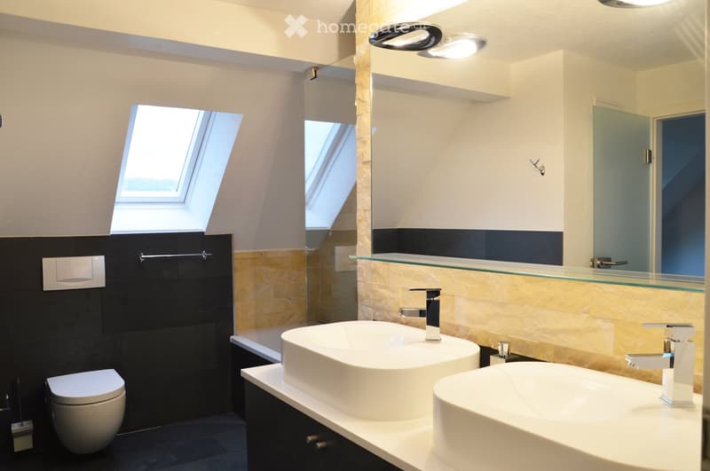 Modern bathroom with two white sinks, a toilet, a bathtub, and a large mirror with a glass shelf above it. The walls are tiled in black and beige, and there is a skylight window providing natural light.