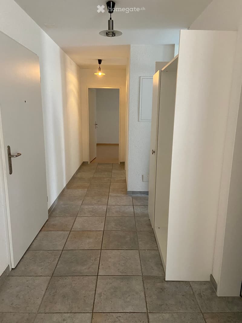 Empty hallway with tiled floor, white walls, and several doors leading to other rooms. Minimal lighting from ceiling fixtures.