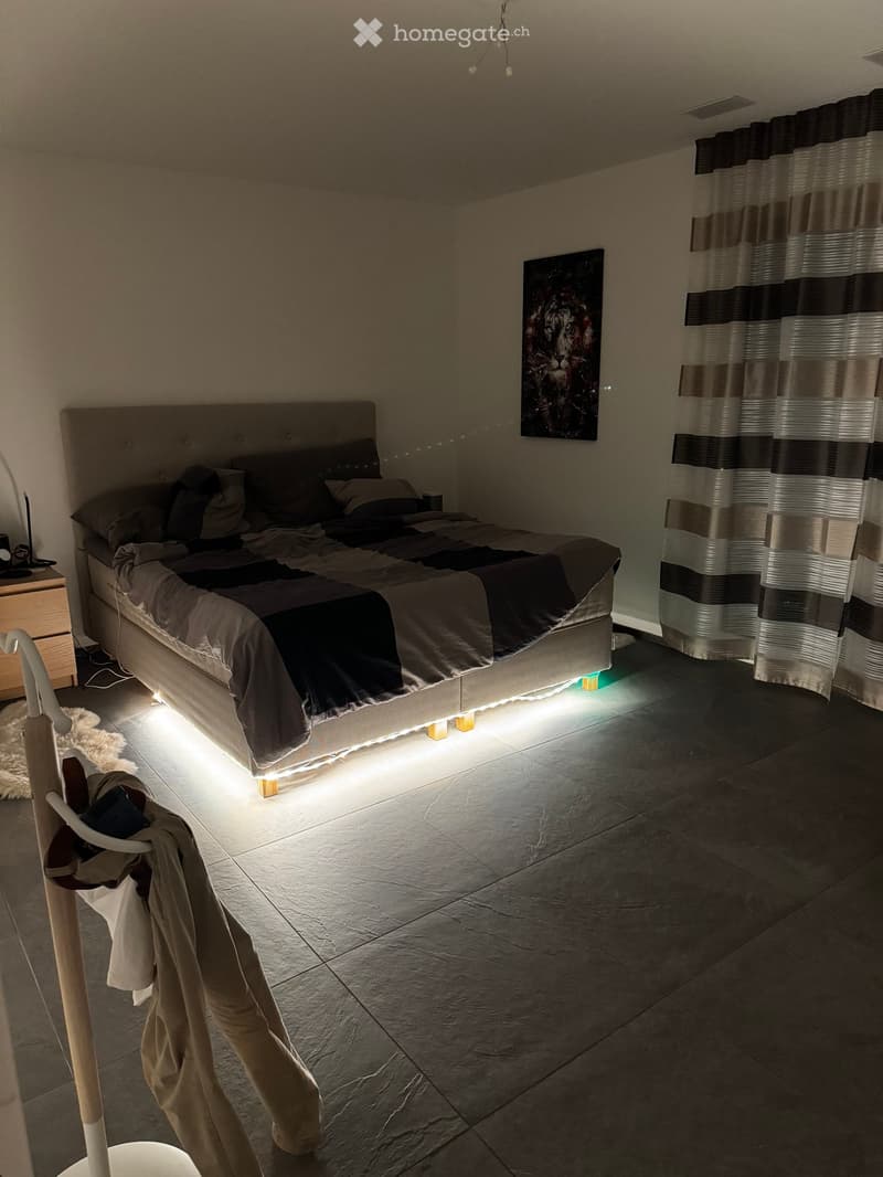 Modern bedroom with a large bed, nightstand, painting, and a curtained window. The room features tiled flooring and ambient lighting under the bed.