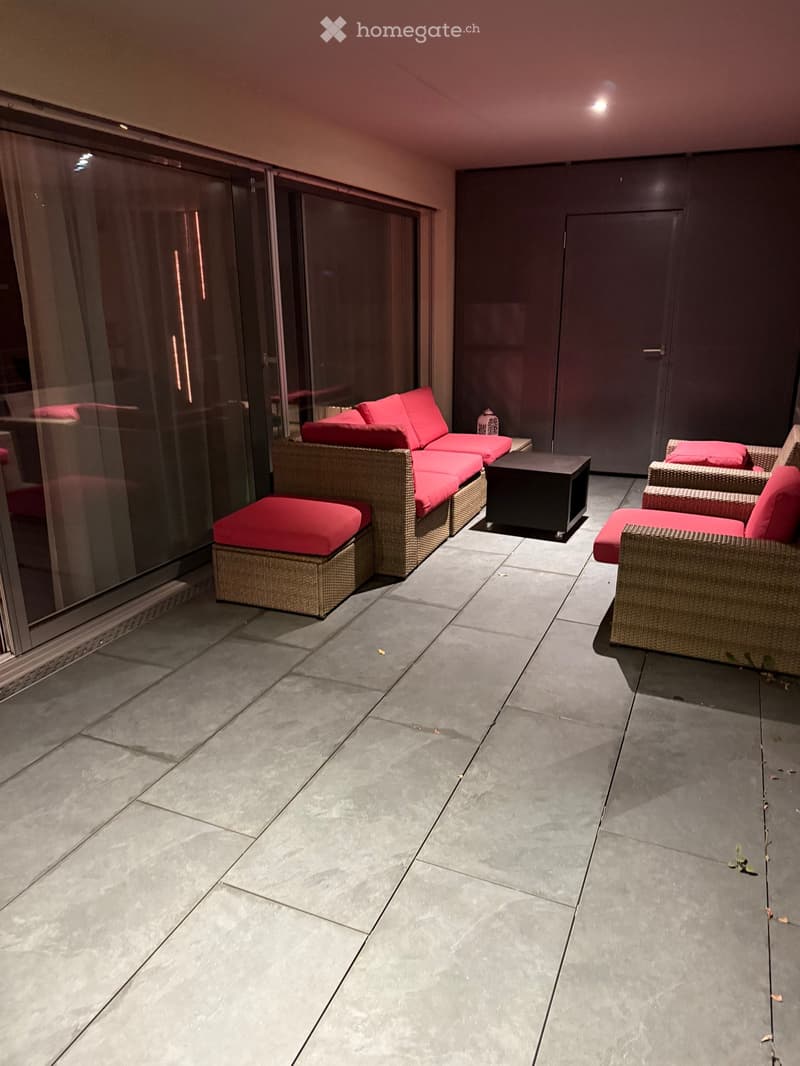 Balcony with grey tiled floor, large glass sliding doors, red-cushioned wicker sofa and chairs, small black side table.