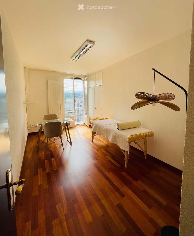 A well-lit room with wooden flooring, a massage table, a glass table with chairs, and a hanging fan.