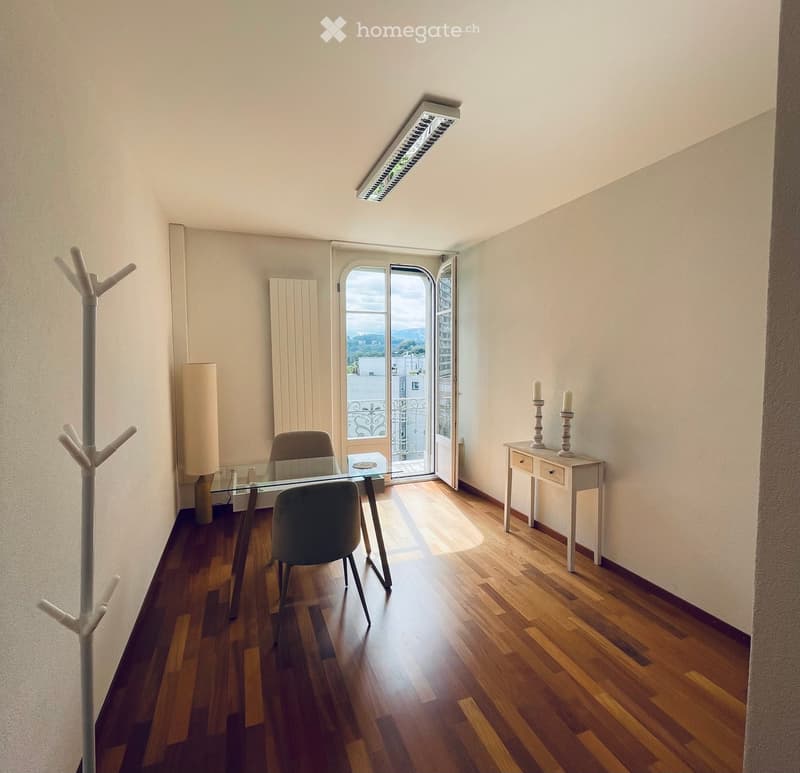Bright, minimalist room with wooden flooring, a glass table, two chairs, a small white table with candles, and a coat rack near the door leading to a balcony with a scenic view.