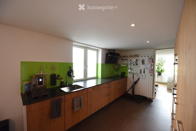 Modern kitchen with wooden cabinets, black countertops, stainless steel sink, coffee machine, and a green accent wall. The space is well-lit with large windows and includes a refrigerator and a vacuum cleaner in the corner.