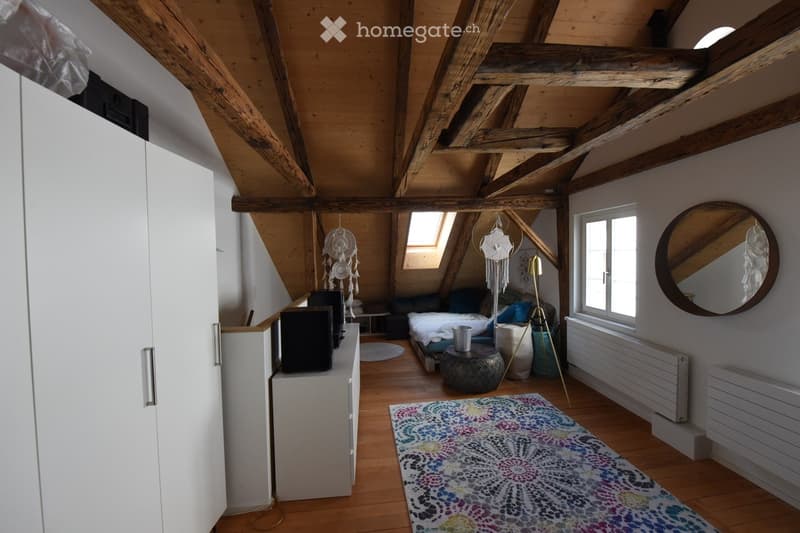 Cozy attic bedroom with wooden beams, skylight, and large window. Features a bed, sofa, small table, mirror, and decorative items.