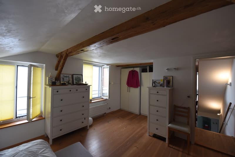 Bright bedroom with wooden floors, exposed wooden beams, large windows with blinds, a bed, a dresser, a wardrobe, a chair, and picture frames on the dresser.