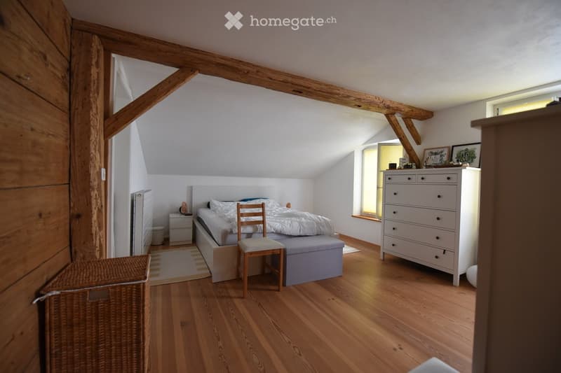 Cozy attic bedroom with wooden beams, hardwood floor, bed, nightstand, dresser, chair, and small stool.