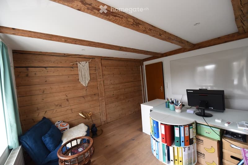 A cozy, well-furnished room with wooden walls and a slanted wooden ceiling. Features include a blue sofa, a small round wooden table, a white desk with a computer monitor, and a colorful file holder. The room also has a decorative woven wall hanging and a
