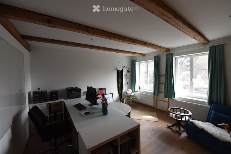 A cozy, well-lit room with wooden beams on the ceiling, a large window with green curtains, a modern desk setup, a comfortable sofa, and a small round table.